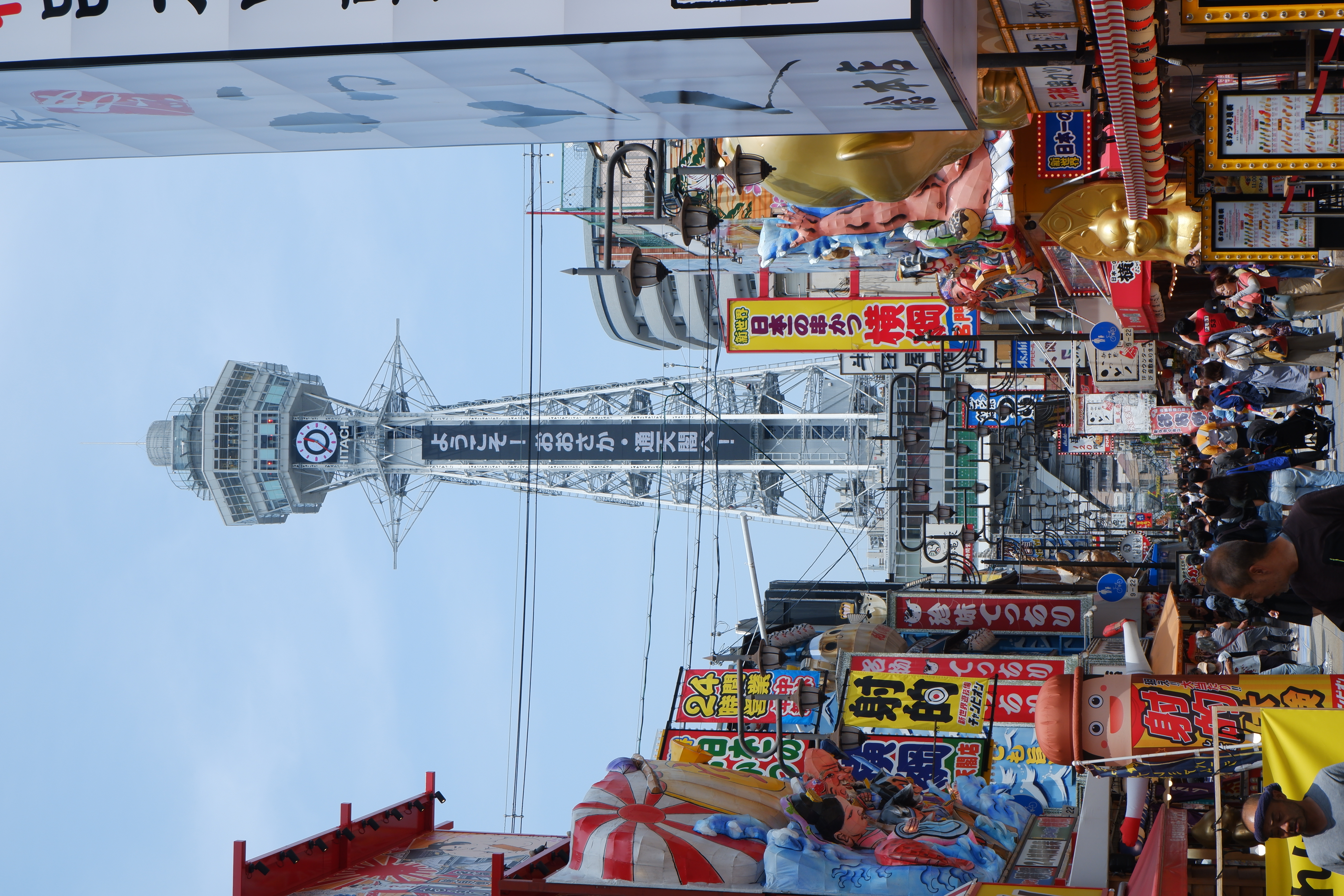 a tower with the word 'Hitachi' at the end of a shopping street